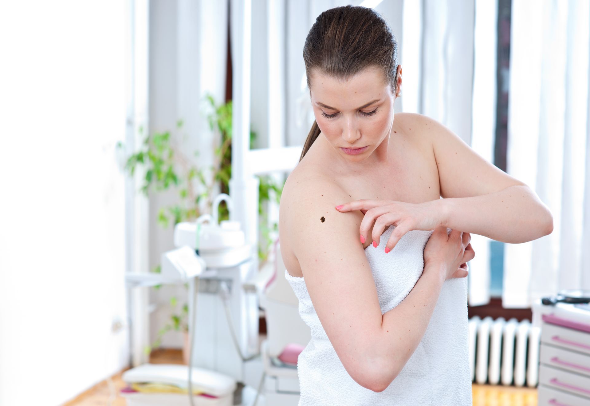 A woman at home in a towel checking a mole on her left shoulder.