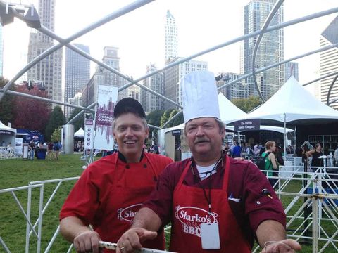 Two chefs in red aprons smiling outdoors at a food event with tents and city buildings behind them