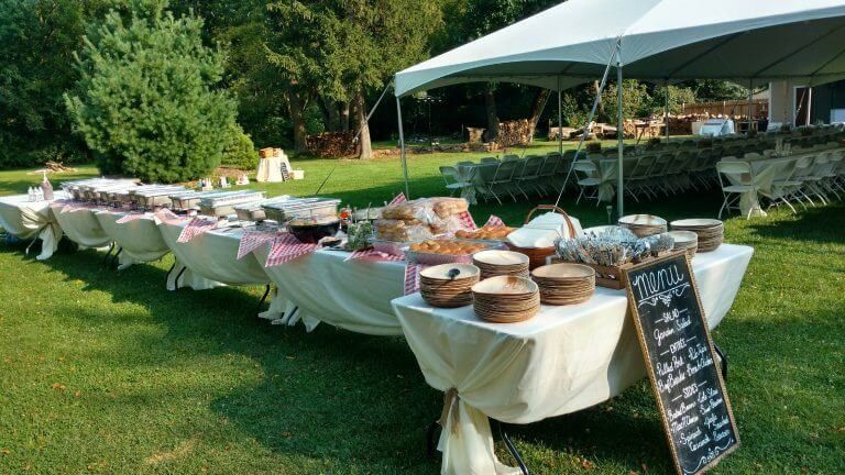 Outdoor buffet tables under a canopy on a grassy lawn, with plates, bowls, and a menu board.