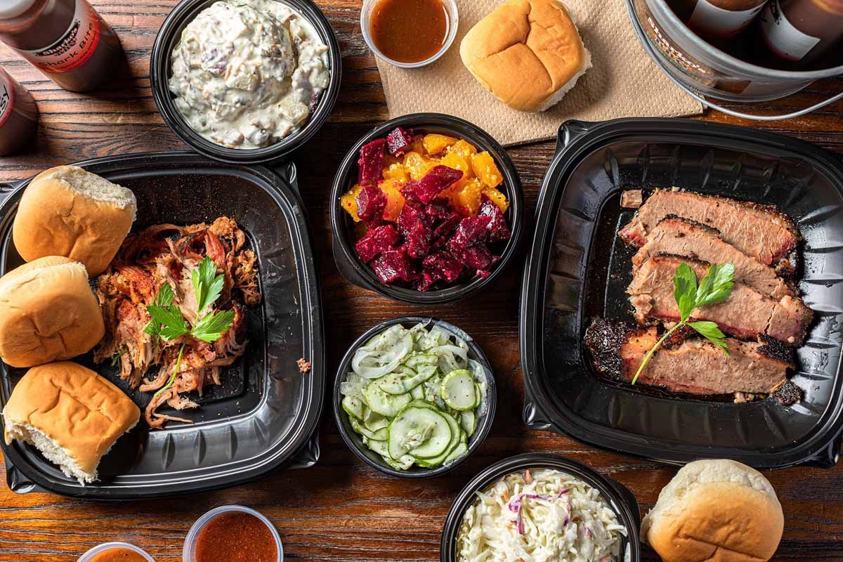 Grilled meat and sliders on trays with sides of coleslaw, pickles, and salad on a wooden table