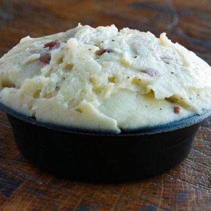 A bowl of creamy mashed potatoes with small bacon bits on a wooden table.