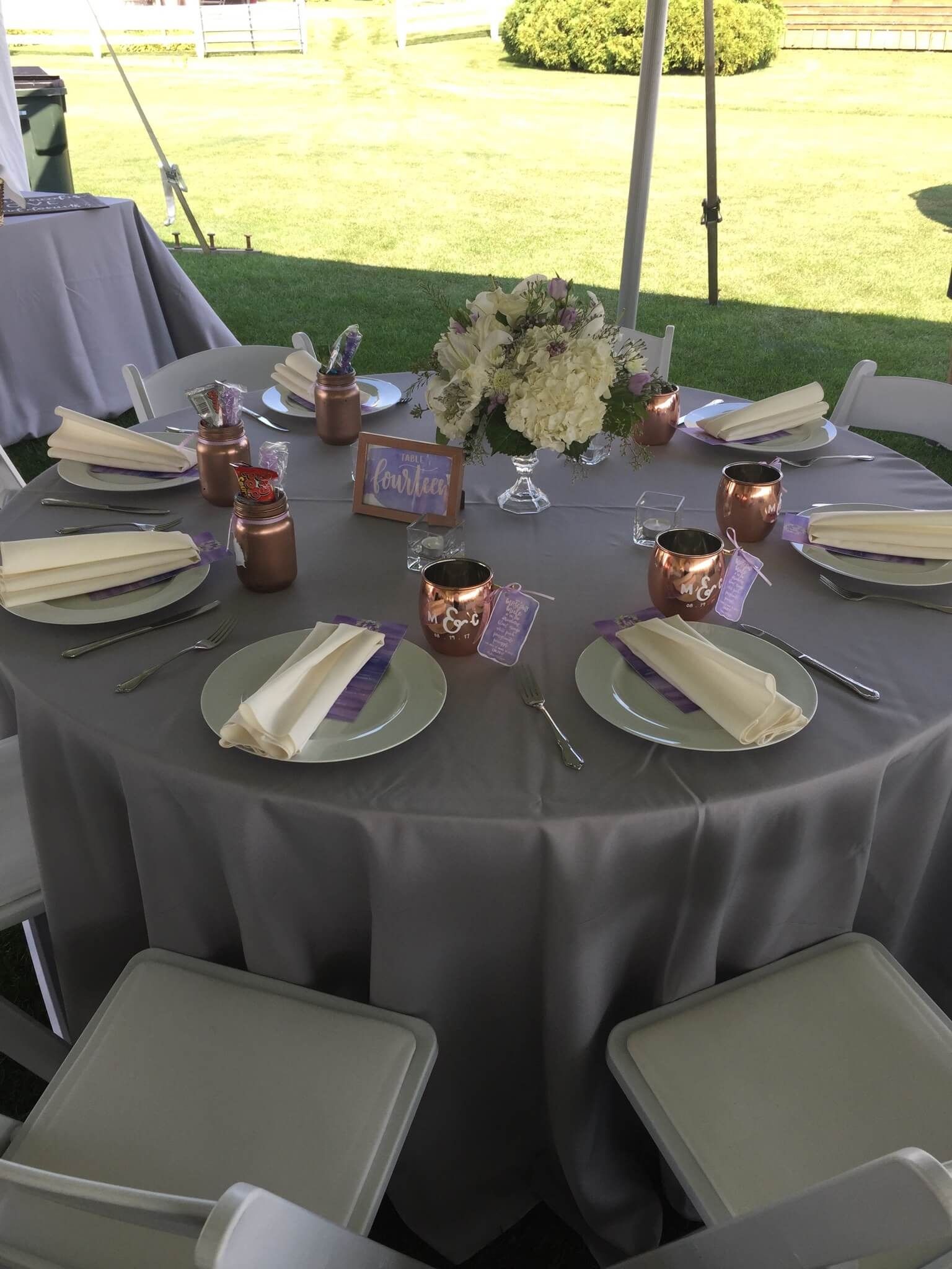 Round outdoor banquet table with gray cloth, plates, candles, and white floral centerpiece under a tent.
