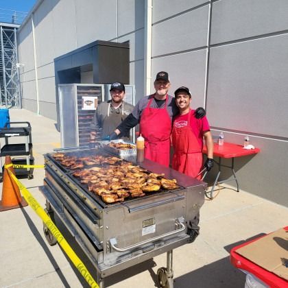 Three people in red aprons grill food outdoors beside a metal building.