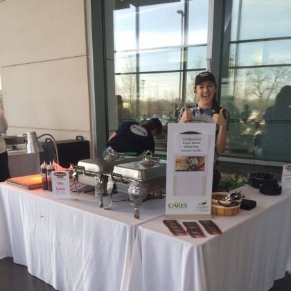 Catering table with trays, dessert cup, and a smiling vendor beside a CARES sign in a glass-walled lobby