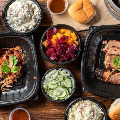 Assorted BBQ meal containers with sides, rolls, and sauces on a wooden table