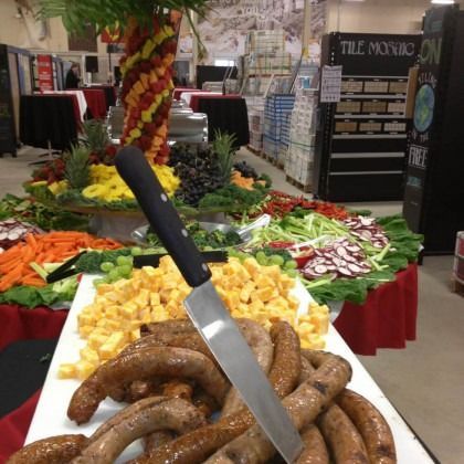 Buffet table with sausage, cheese cubes, vegetables, and a knife in the foreground at a grocery store.