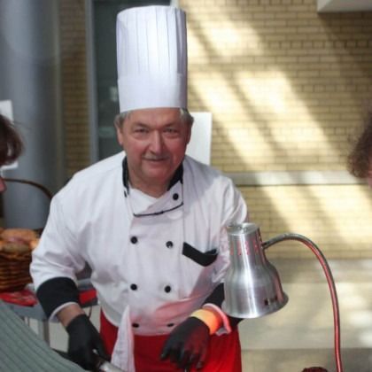 Chef in white uniform and tall hat cooking at a counter with a heat lamp.