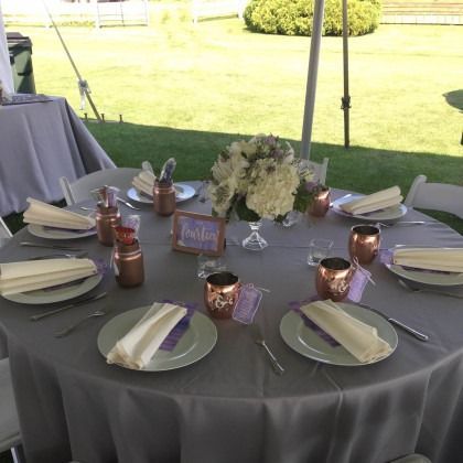 Round outdoor banquet table with gray cloth, white plates, folded napkins, candles, and a floral centerpiece under a tent.