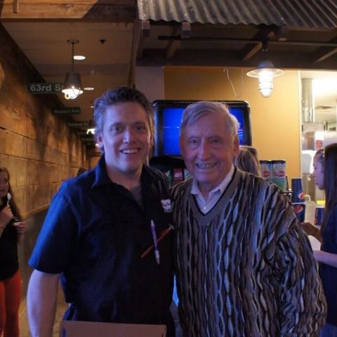 Two men smiling and posing together indoors under warm lights, one in a black polo and one in a striped sweater