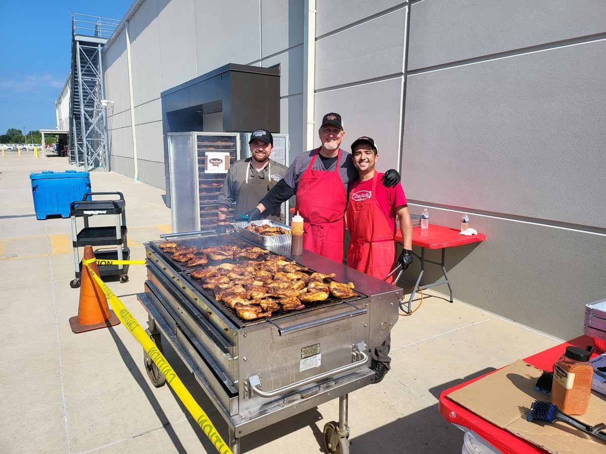 Three people in red aprons grilling food on a large outdoor barbecue beside a building.
