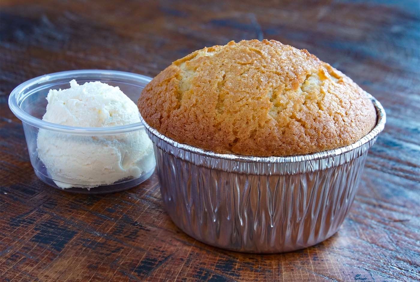 Golden muffin in a foil cup beside a small bowl of white frosting on a wooden table