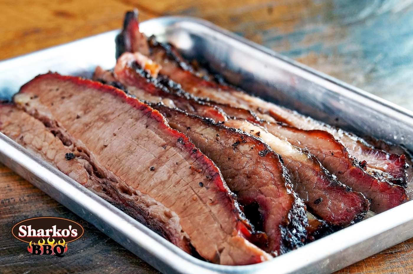 Sliced smoked brisket in a metal tray, showing a dark bark and pink smoke ring