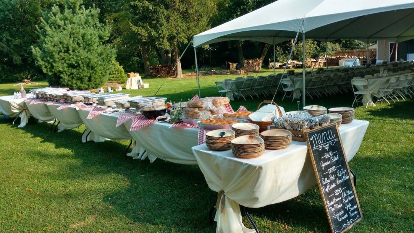 Outdoor buffet table with covered dishes and desserts on white cloths under a tent on a grassy lawn