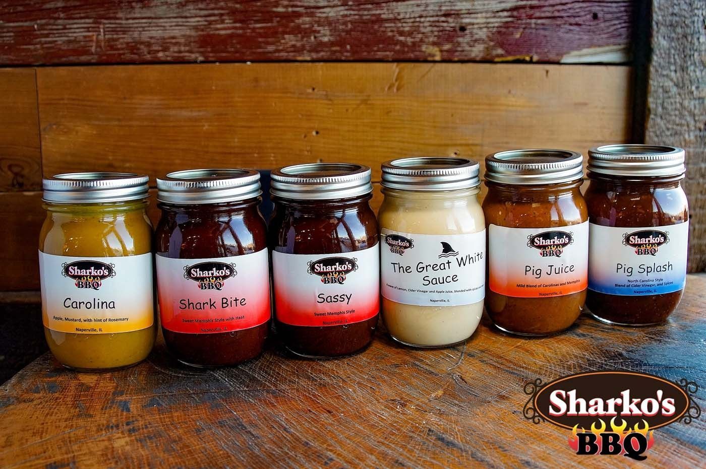 Six jars of colorful barbecue sauce lined up on a wooden table with a rustic wood backdrop.