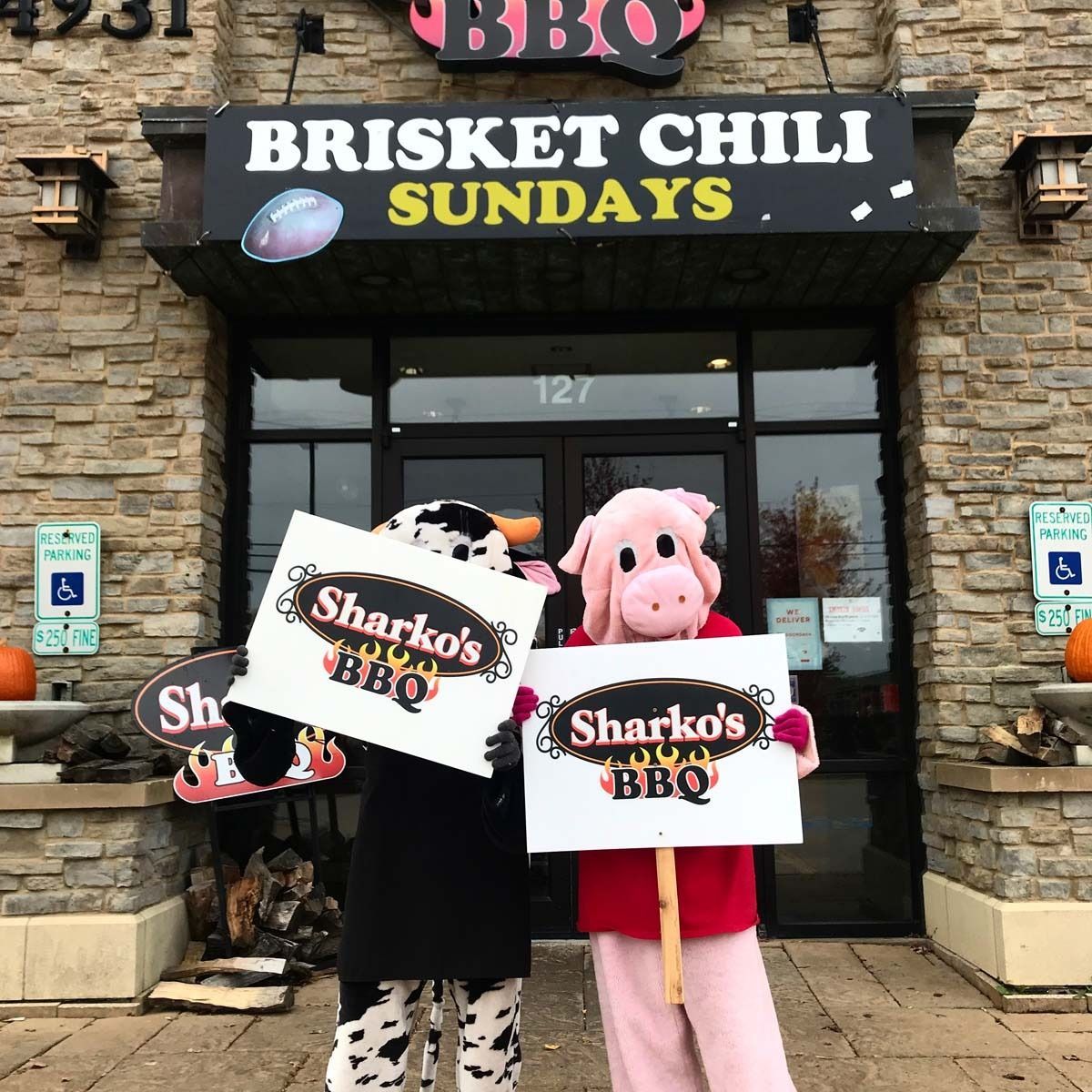 Two people in pig costumes hold Sharko’s BBQ signs outside a restaurant under a Brisket Chili Sundays banner