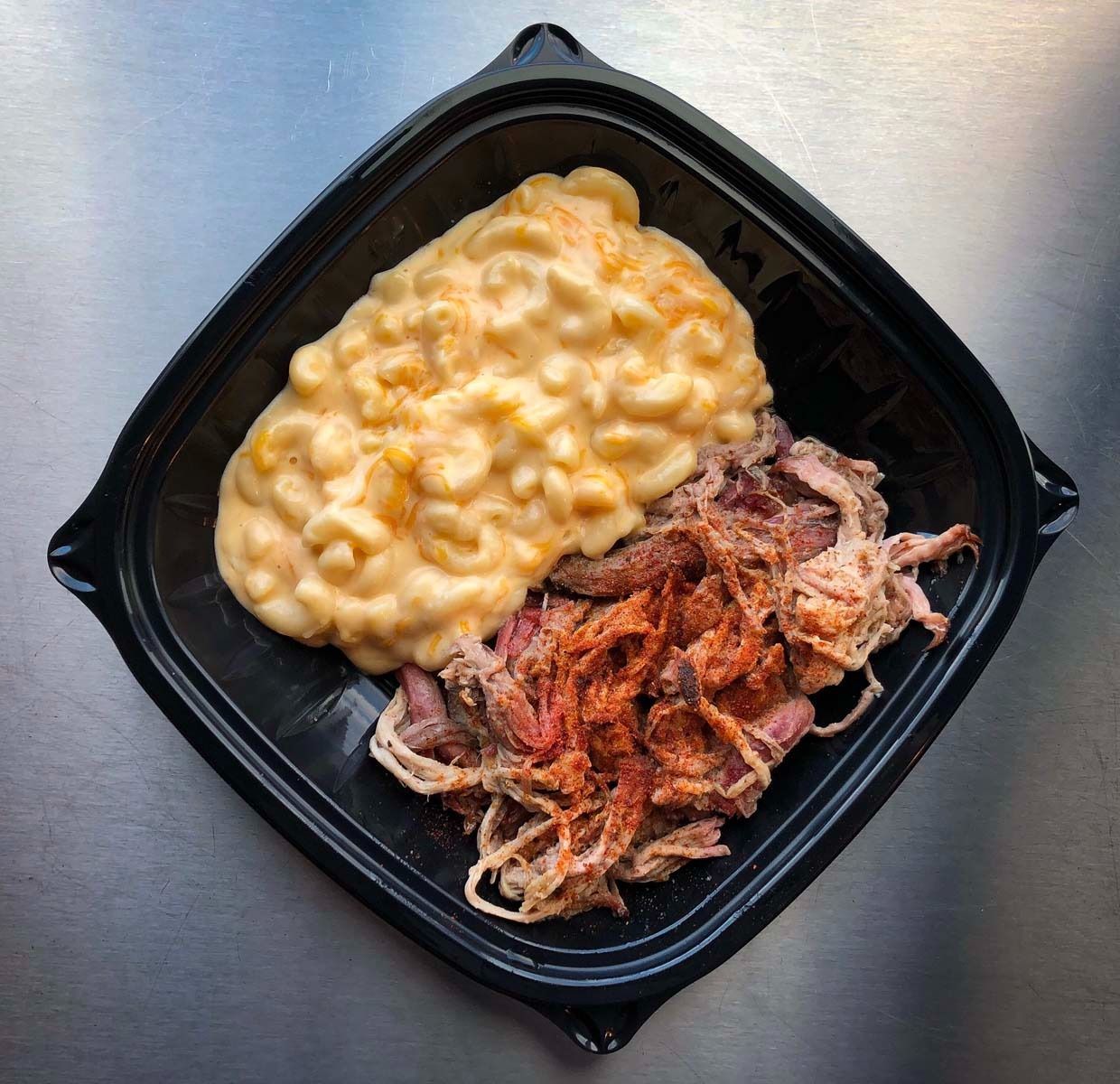 Black takeout tray with creamy macaroni and cheese and shredded pulled pork on a metal surface