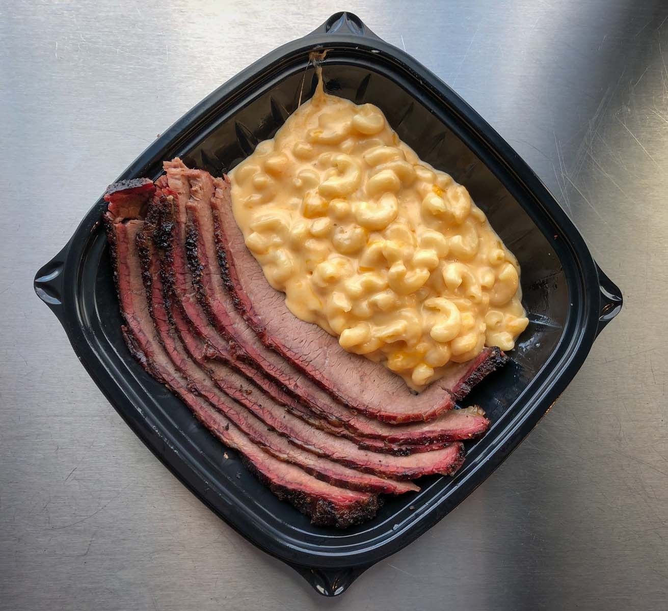 Sliced brisket and macaroni and cheese in a black takeout container on a metal table.