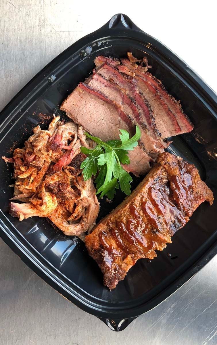 Grilled meat platter with sliced brisket, ribs, and pulled pork on a black tray, garnished with parsley.