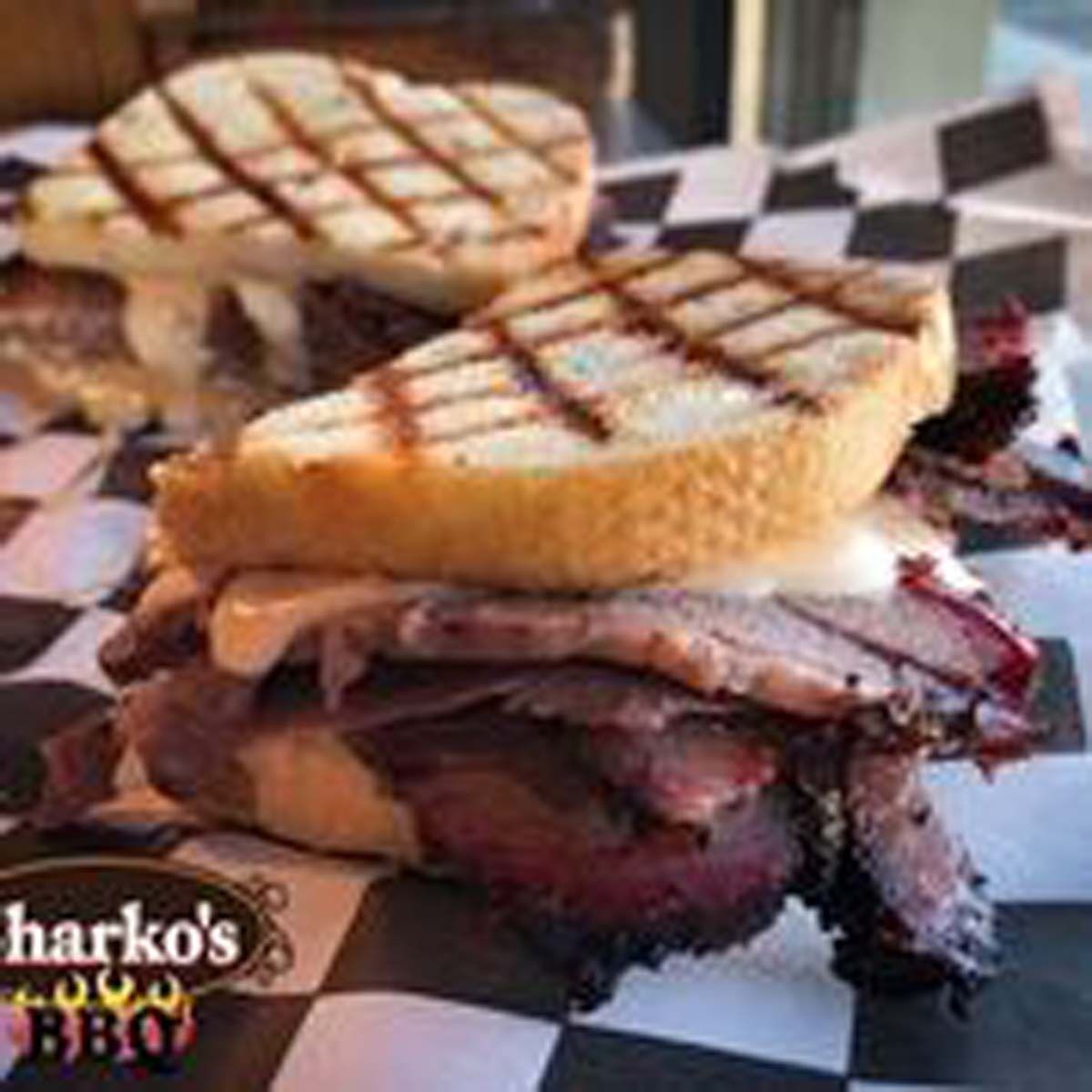 Grilled sandwich with melted cheese and bacon on checkered paper, with a BBQ logo in the corner