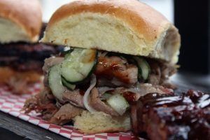Close-up of a pulled pork sandwich with pickles on a tray, with barbecue sauce beside it.
