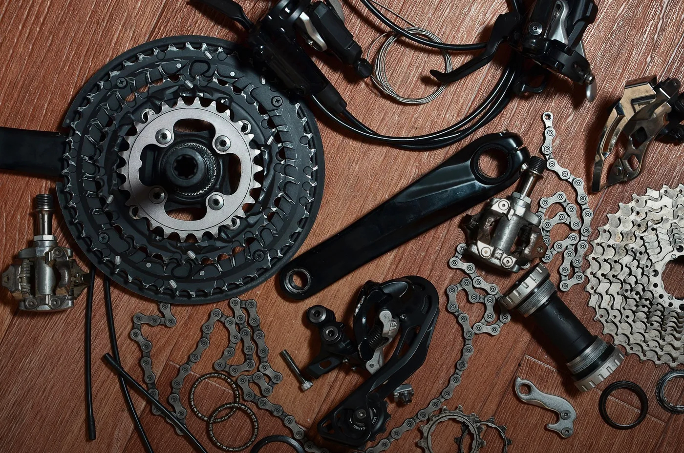 Bicycle drivetrain components: chainrings, cassette, derailleur, crankset, pedals, cables, and chain, scattered on a wood surface.