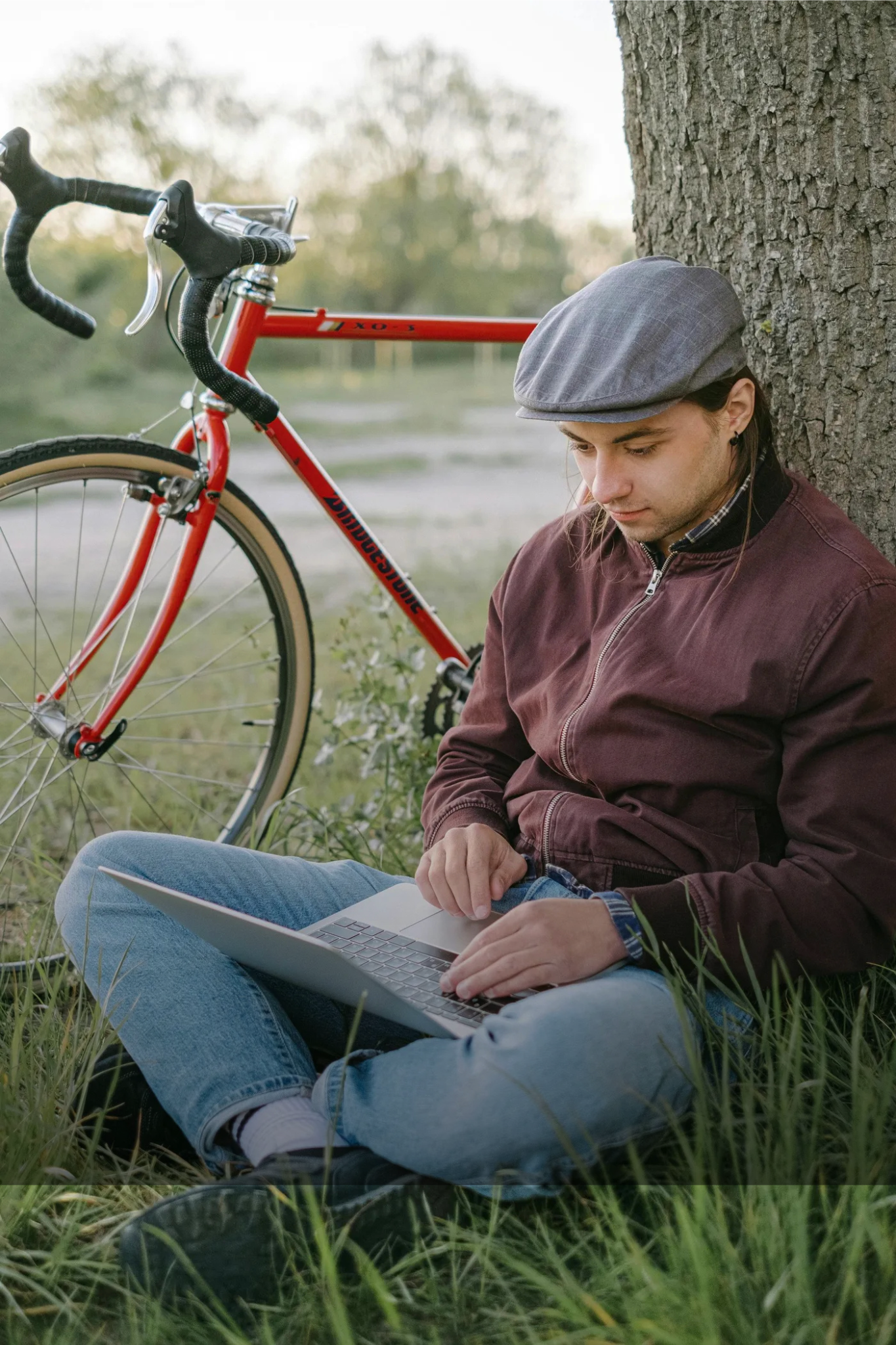 Man sitting against a tree, working on a laptop, with a red bicycle beside him.