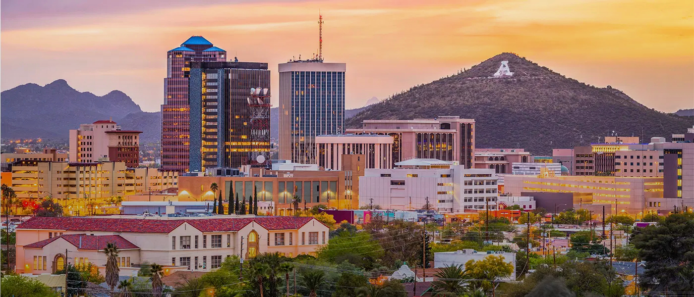 City skyline at sunset with a mountain, illuminated buildings, and pink and yellow sky.