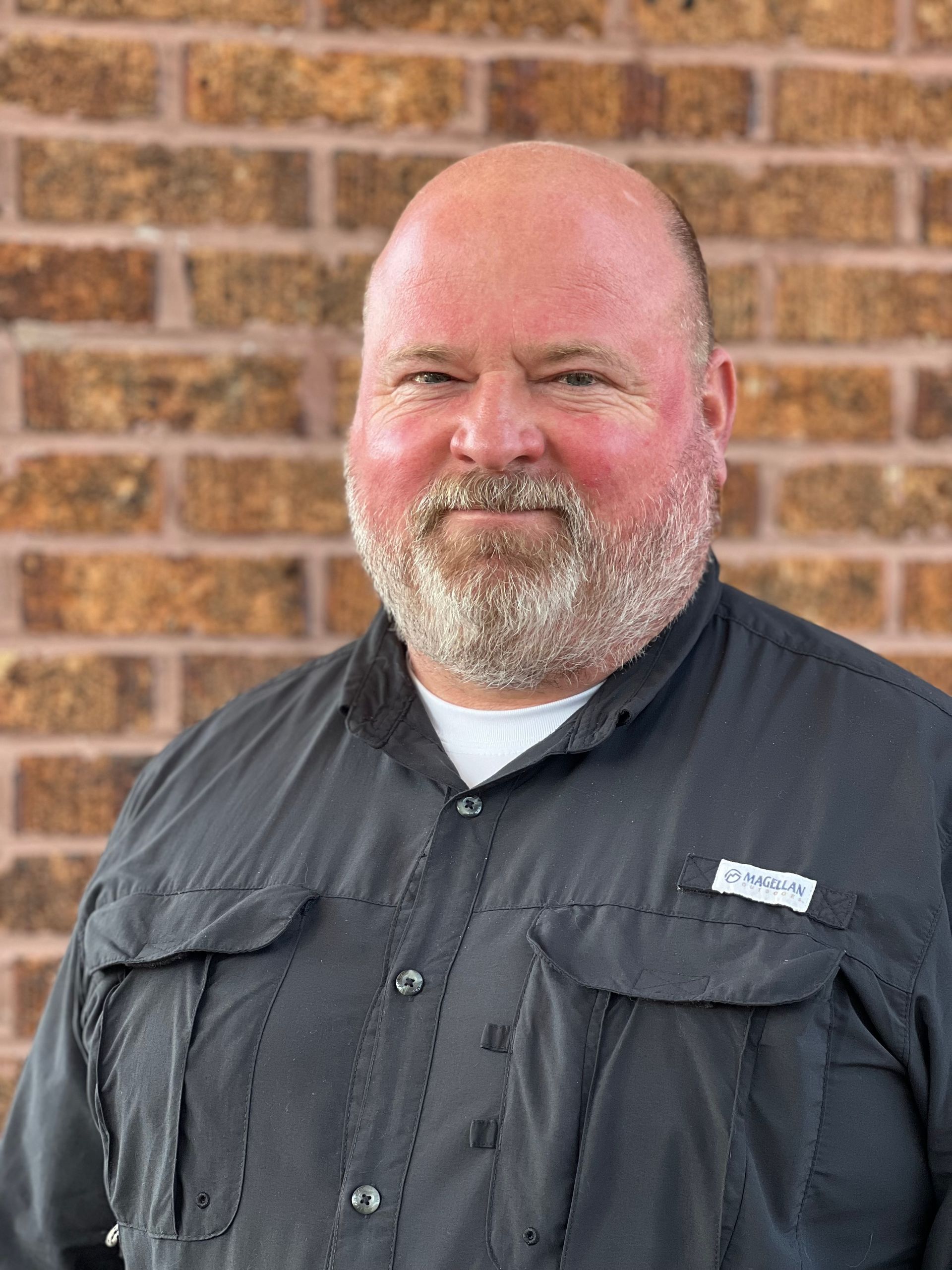 Smiling bald man with a gray beard in a dark shirt standing before a brick wall
