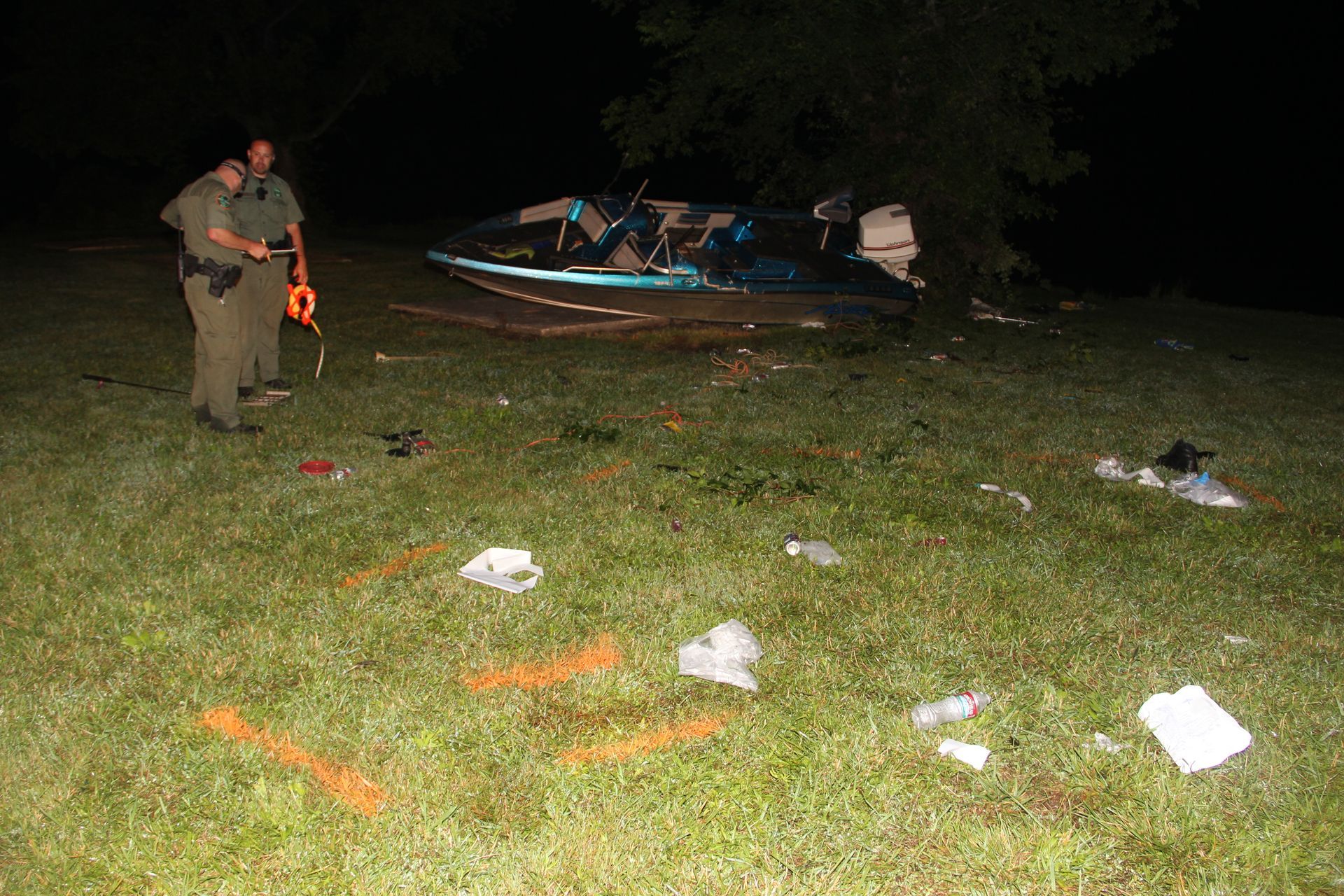 Police officer near a boat at night, with evidence markers scattered on grassy ground.