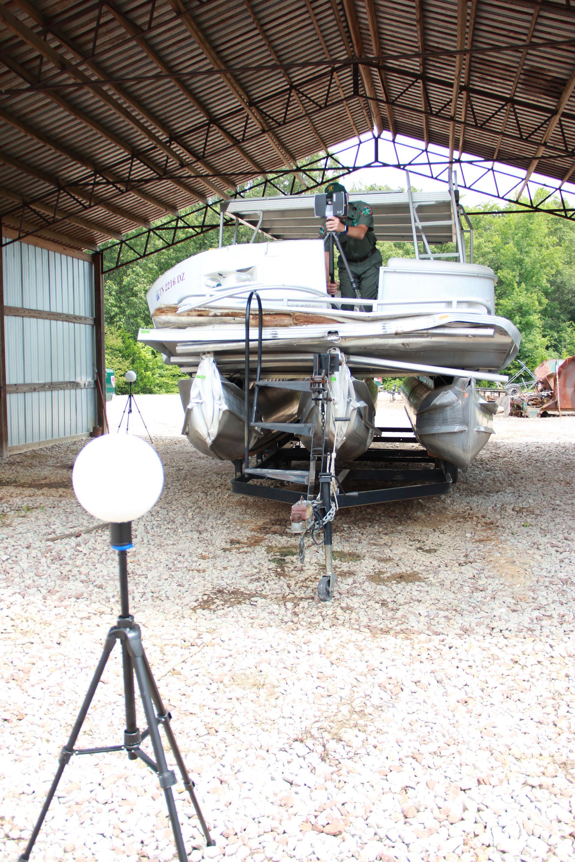 White pontoon boat on a trailer in a covered shed, with a tripod-mounted spherical device in the foreground.