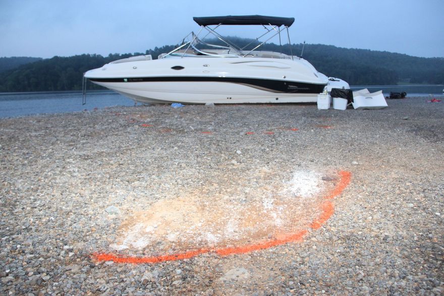 White speedboat on a rocky beach with an orange survey mark in the foreground, water and trees behind.