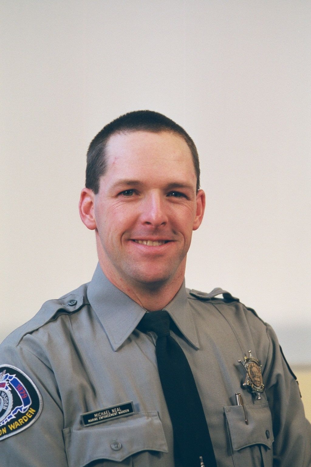 Smiling uniformed police officer in gray shirt and black tie against a plain background