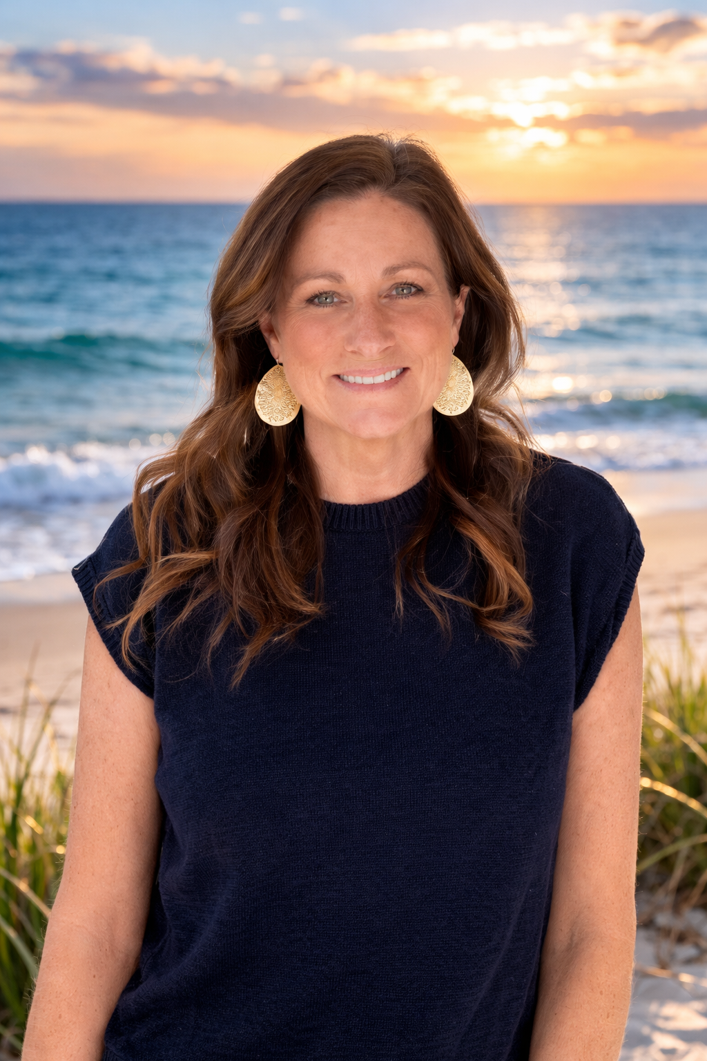 Woman smiling on a beach at sunset, with ocean waves and warm sky behind her.