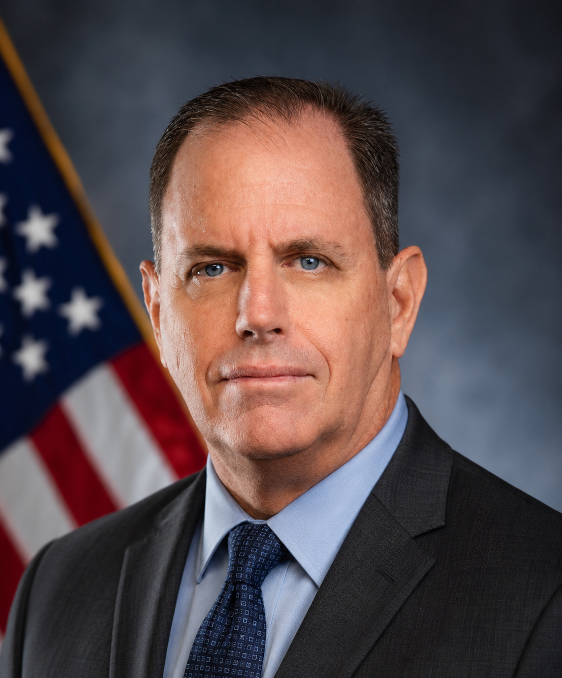 Formal portrait of a man in a suit and tie, with a U.S. flag blurred in the background