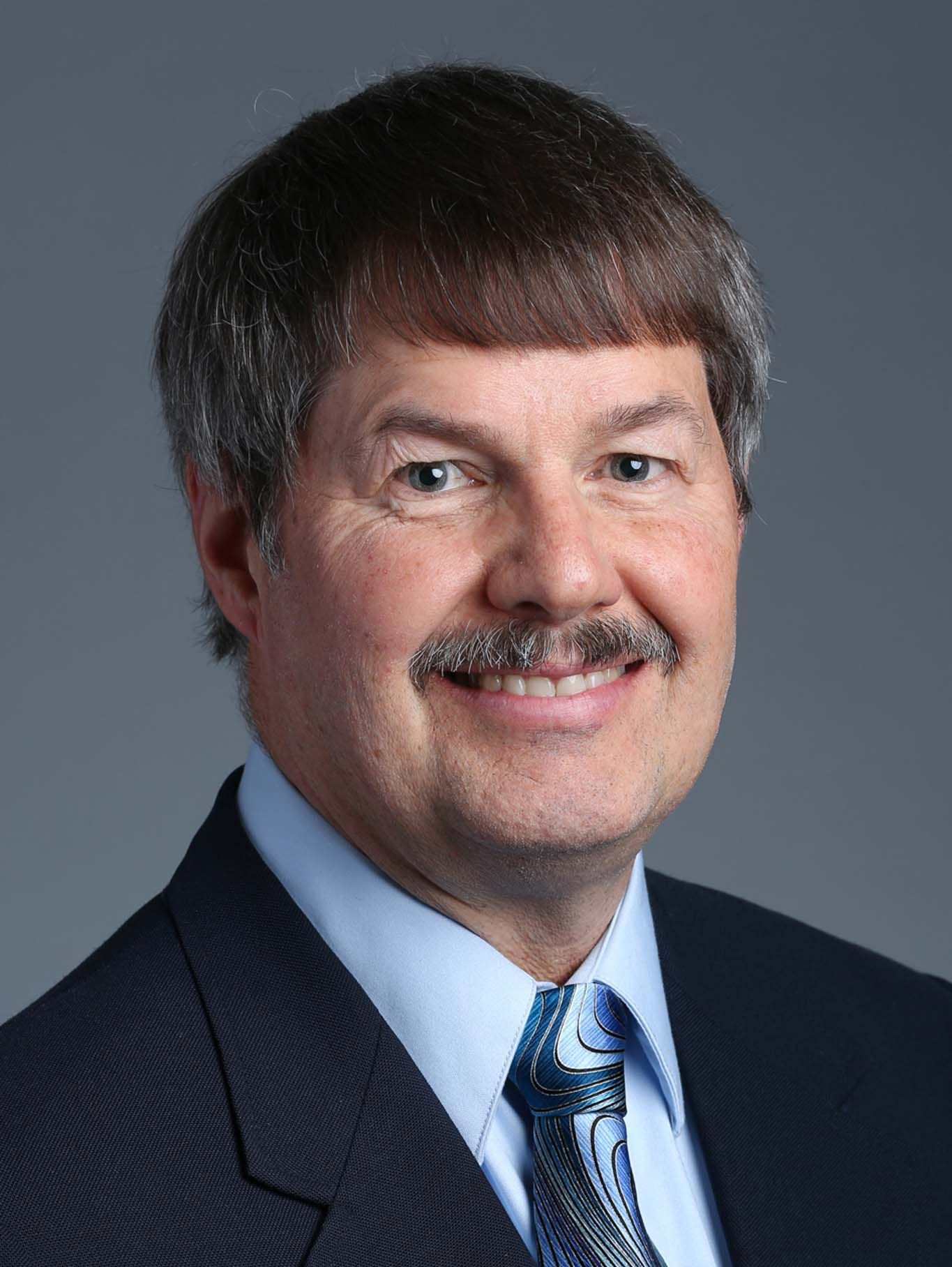 Portrait of a smiling man in a dark suit, light blue shirt, and patterned tie on a gray background
