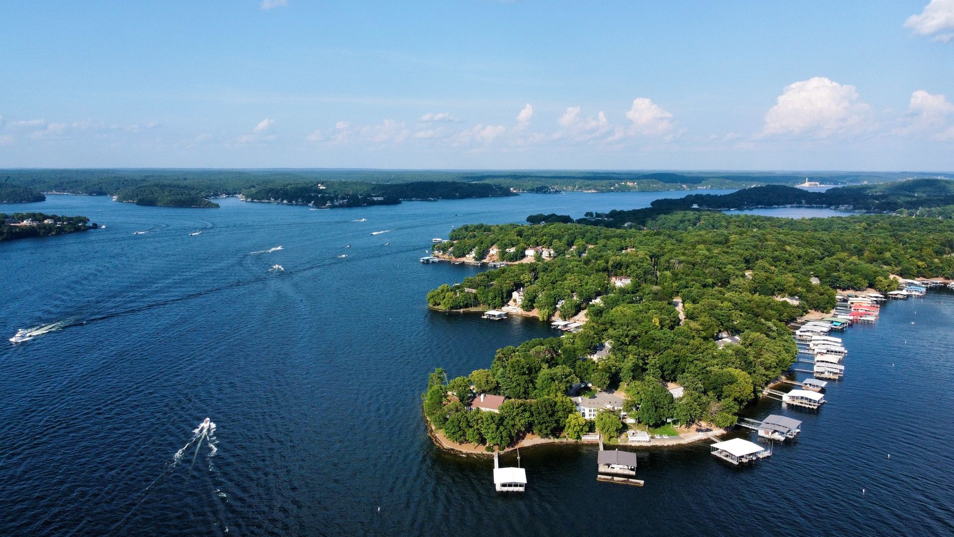 Aerial view of a blue lake with green islands and boats docked along a wooded shoreline.