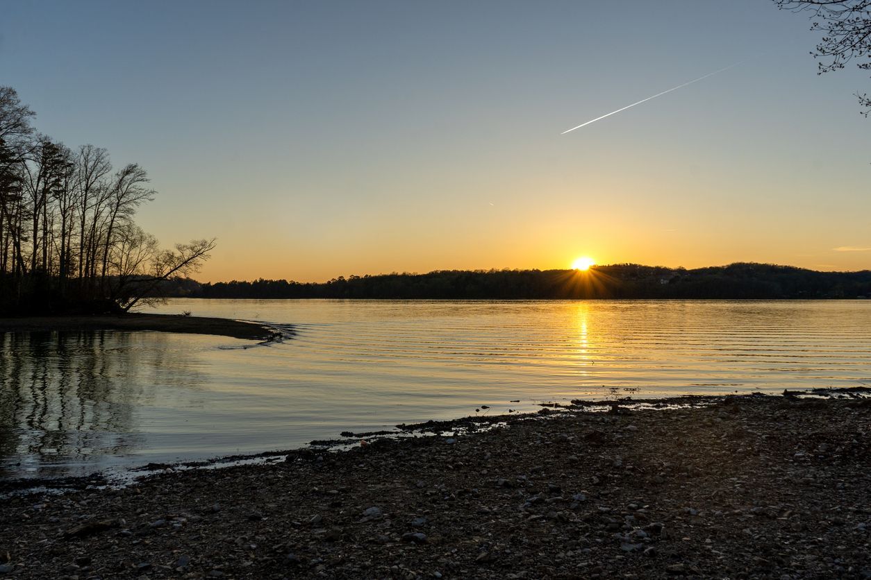 Sunset over a calm lake with silhouetted trees and a rocky shore