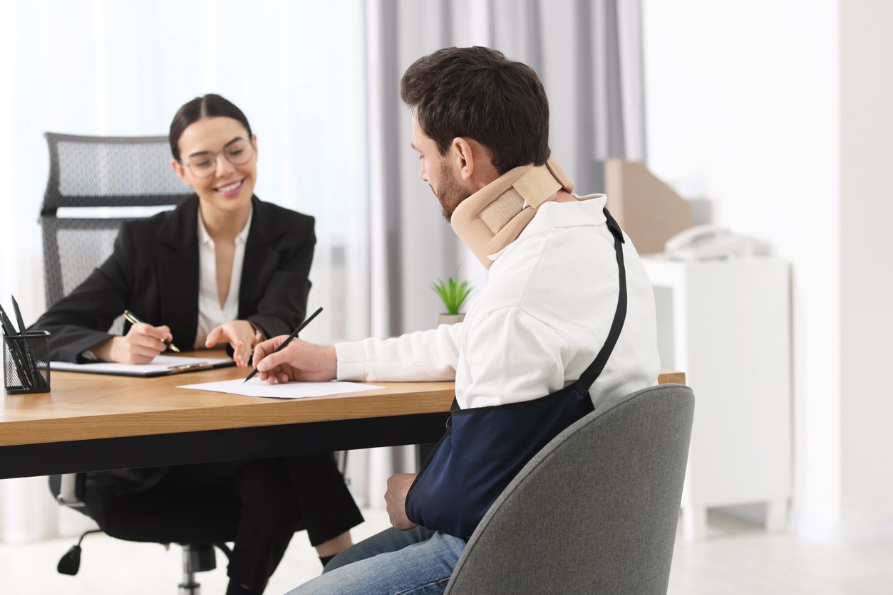 Two people talking at a table in a bright office, one with a neck brace and arm sling