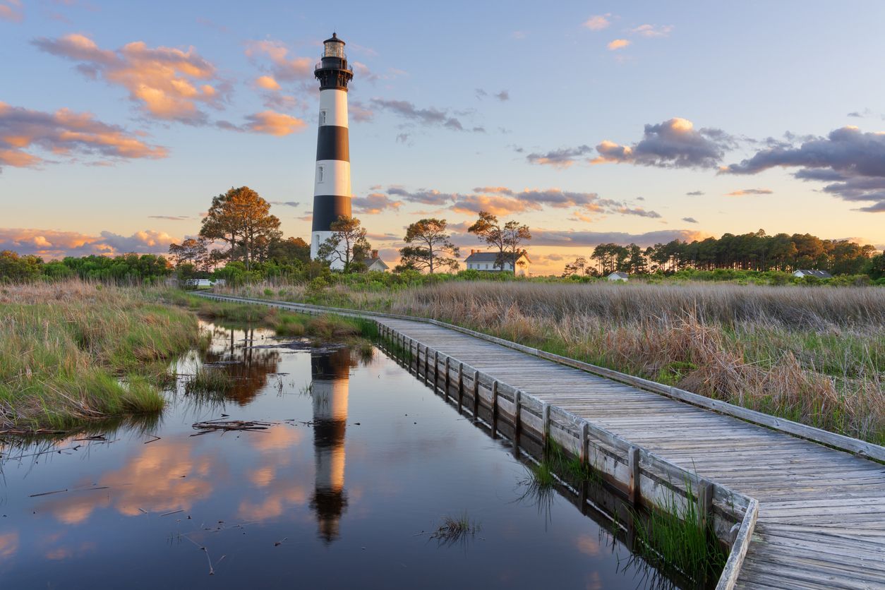 Lighthouse at sunset beside a marsh, with a wooden boardwalk and reflection in calm water.