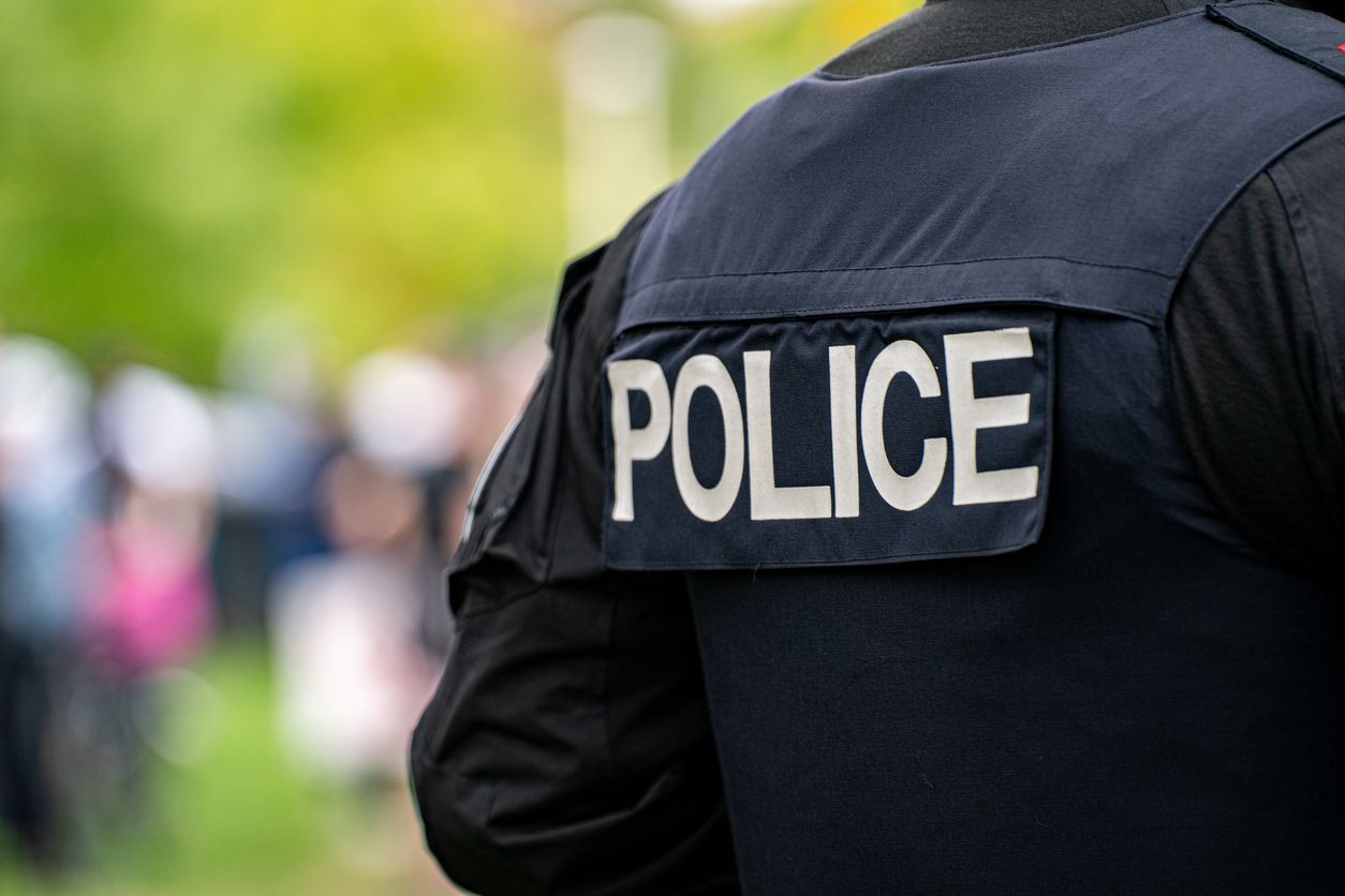 Police officer seen from behind in black uniform with “POLICE” patch outdoors, blurred crowd in background