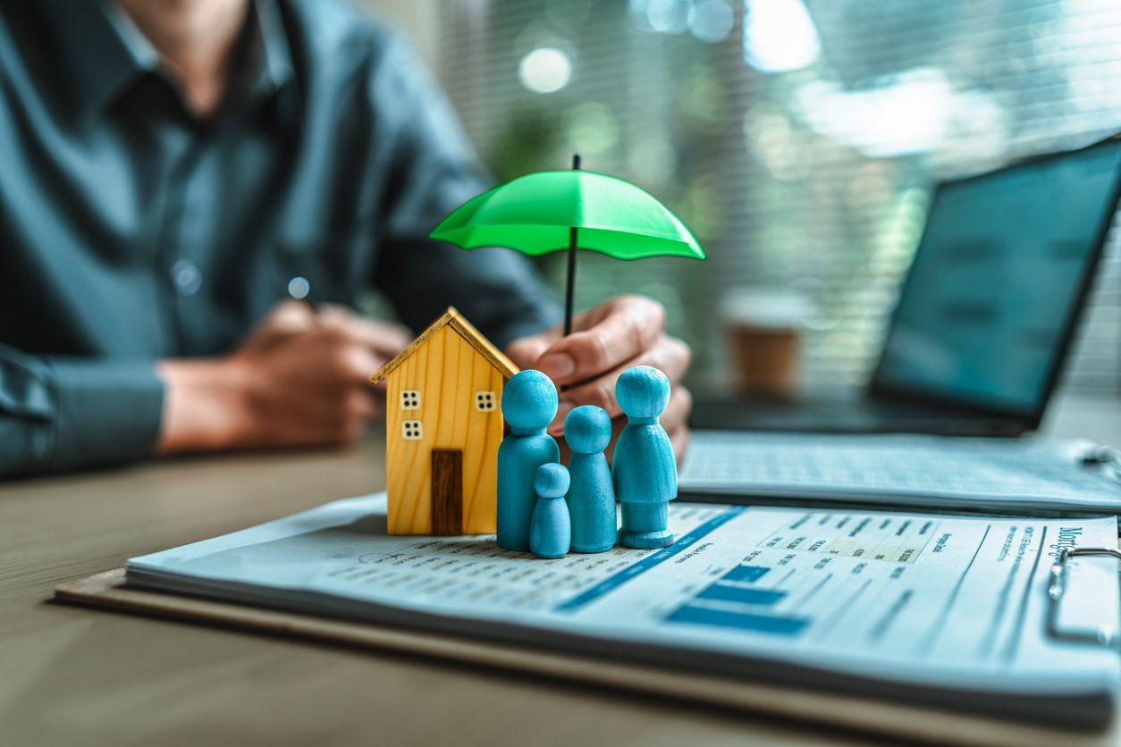 Small blue family figurines beside a yellow house and green umbrella on papers, with a laptop in the background