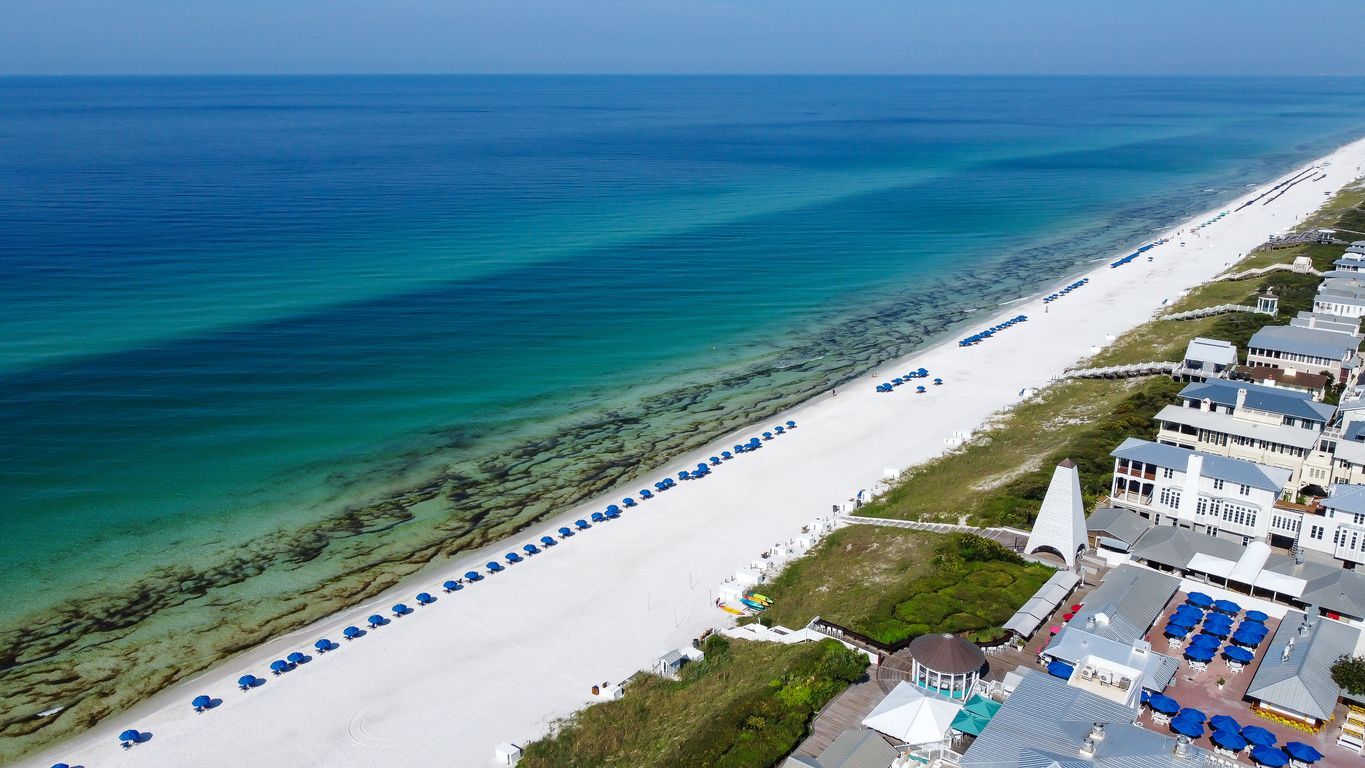 Aerial view of a white sandy beach with turquoise water and rows of blue umbrellas beside resort buildings