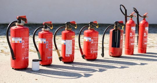 Estintori rossi allineati su una superficie di cemento, con un piccolo contenitore bianco.