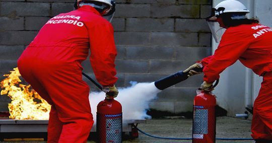 Due persone in tute rosse e caschi usano gli estintori per spegnere un piccolo incendio.