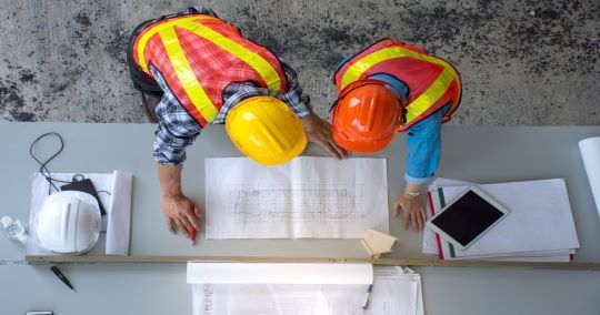 Due operai edili, con indosso giubbotti di sicurezza e caschi protettivi, esaminano i progetti seduti a un tavolo.