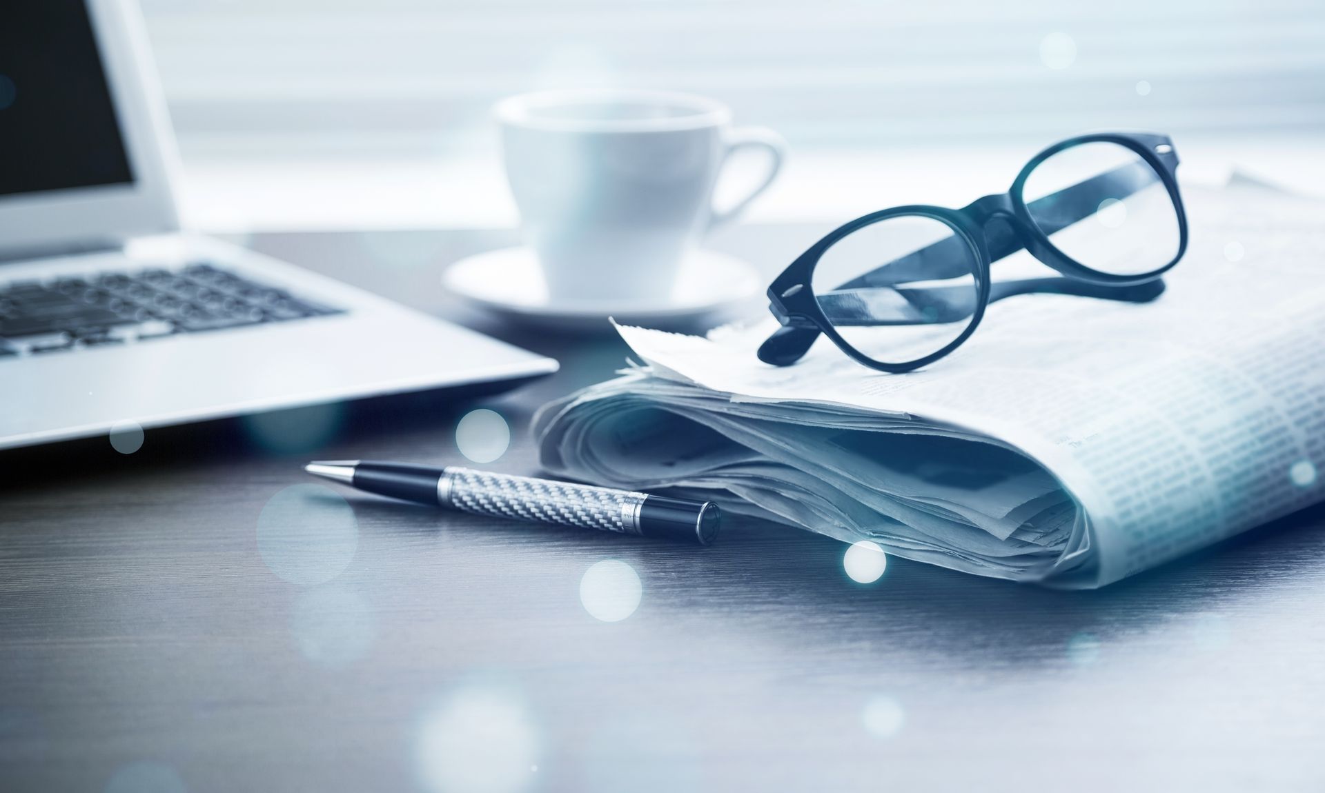 Computer portatile, occhiali, giornale, penna e tazza di caffè su una scrivania.