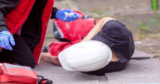 Persona ferita a terra, che riceve il primo soccorso. Un medico con giacca rossa e guanti è inginocchiato lì vicino.
