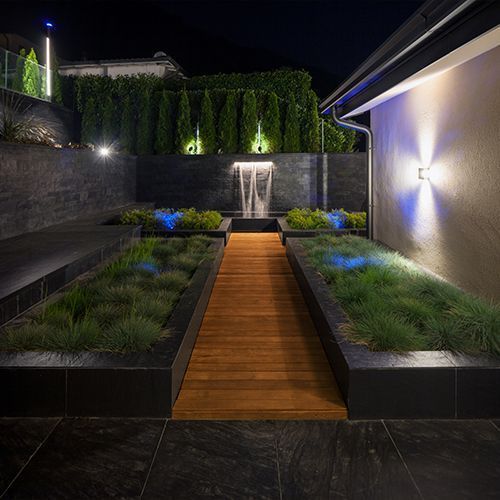 Night landscape view with a wooden walkway between grassy beds, a fountain, and lighting in Crosby