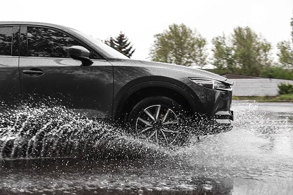 Gray SUV splashing through a puddle on a wet road, trees in the background. | Dave's Automotive LLC