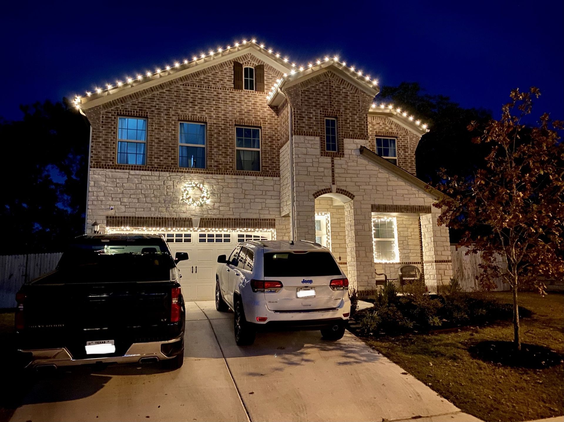 A two-story house at night with white Christmas lights. Cars sit in the driveway.